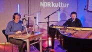 Links sitzt ein mann vor einem PC und rechts neben ihm sitzt ein Mann an einem schwarzen Flügel, sie machen gemeinsam Musik. © Screenshot / NDR Foto: Screenshot / NDR