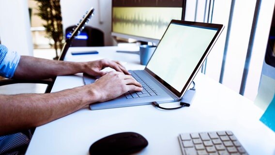 Männerhände tippen etwas in eine Laptop-Tastatur. Im Hintergrund eine Gitarre und ein weiter Bildschirm. © IMAGO / Westend61 