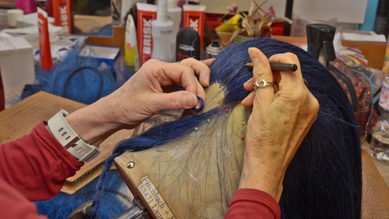 Eine Künstlerin arbeitet Haare in eine Perücke ein und hält dafür eine gekrümmte Nadel in der Hand, die jedes blaue Haar in eine Masche hakt © NDR Foto: Patricia Batlle