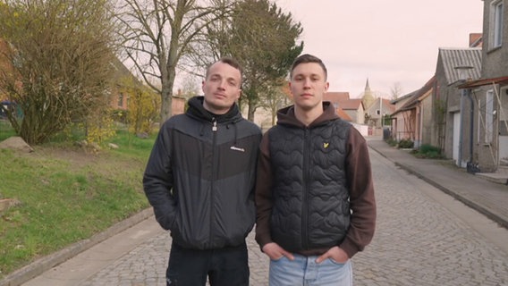 Zwei Männer in dunklen Jacken stehen auf einer Straße und schauen in die Kamera © Screenshot NDR 