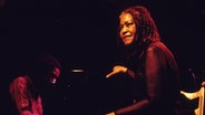 Die amerikanisch Sängerin Abbey Lincoln sitzt bei einem Konzert in Hamburg auf der Bühne und lächelt ins Publikum. © picture alliance / Jazzarchiv Foto: Jazzarchiv / Hogreve