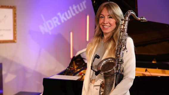 Eine Frau mit langen blonden Haaren steht vor einem Flügel und hat ein Saxophon untern Arm, dabei lacht sie. © Tim Piotraschke / NDR Foto: Tim Piotraschke / NDR