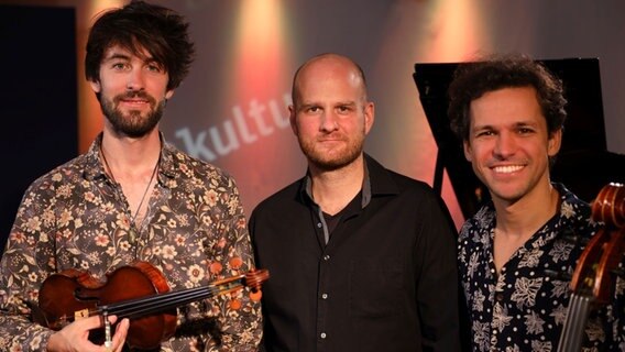 Drei Männer stehen einander, ganz links der Mann hält eine Geige in der Hand und rechts der Mann stützt sich am Cello ab. © Franziska Dieckmann / NDR Foto: Franziska Dieckmann / NDR