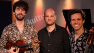 Drei Männer stehen einander, ganz links der Mann hält eine Geige in der Hand und rechts der Mann stützt sich am Cello ab. © Franziska Dieckmann / NDR Foto: Franziska Dieckmann / NDR