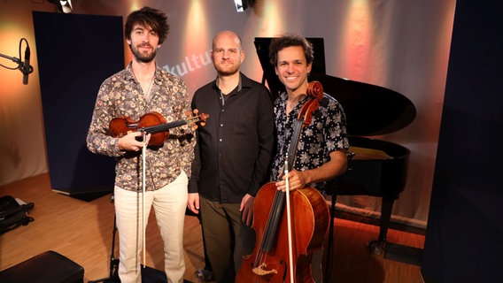 Drei Männer stehen einander, ganz links der Mann hält eine Geige in der Hand und rechts der Mann ein Cello. © Franziska Dieckmann / NDR Foto: Franziska Dieckmann / NDR
