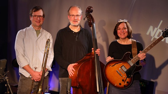 Drei Leute stehen nebeneinander und halten von links eine Posaune, einen Kontrabass und eine E-Gitarre in der Hand, zwei Männer links und eine Frau rechts. © Tim Piotraschke / NDR Foto: Tim Piotraschke / NDR