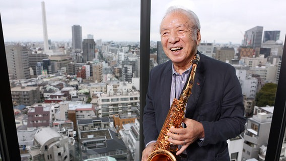 Ein Mann posiert mit Altsaxofon vor einem Fenster. Im Hintergrund ist die Stadt Tokio zu sehen. © Toshiyuki Kon / picture alliance Foto: Toshiyuki Kon