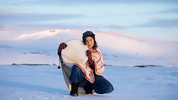 Eine dunkelhaarige Frau mit Mütze und weiß-rotem Cape kniet in einer Schneelandschaft und hält eine Rahmentrommel. © Nordischer Klang Foto: Elisabeth Jøraholmen
