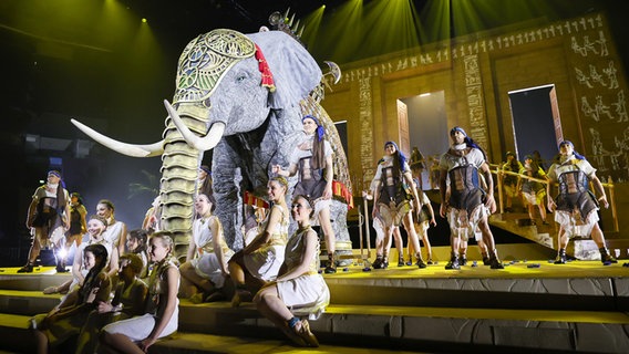 Darstellerinnen und Darsteller stehen mit einem Elefanten auf der Bühne in der Barclays-Arena Hamburg nach einer Pressekonferenz zum Opern-Spektakel "Aida" von Giuseppe Verdi. © picture alliance/dpa | Christian Charisius 