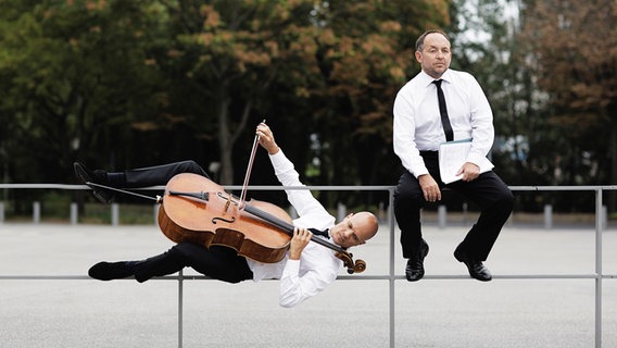 Zwei Männer: einer sitzt auf einem Geländer, der andere hängt daran und spielt dabei Cello. Zwei Männer: einer sitzt auf einem Geländer, der andere hängt daran und spielt dabei Cello. © Nikolaj Lund Foto: Nikolaj Lund