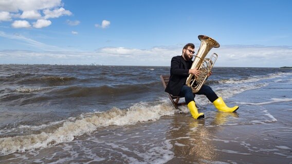 Ein Mann mit gelben Gummistiefeln sitzt im Wasser am Meer und spielt Tuba © Karlheinz Kraemer Foto: Karlheinz Kraemer