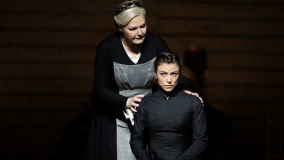Das Bild zeigt zwei weibliche Personen. Eine Frau steht hinter einer anderen sitzenden Frau. Die stehende Person hat helles Haar, welches nach hinten dunkler wird, und trägt ein schwarzes Kleid mit einer hellgrau gestreiften Schürze. Die sitzende Frau hat dunkles Haar und trägt ein dunkles Kleid. © Camilla Greenwell 