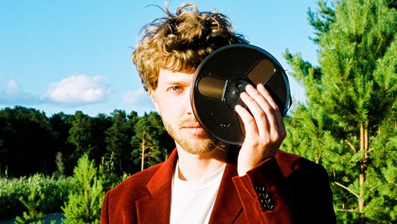 Ein junger Mann schaut in die Kamera und hält dabei das linke Auge mit einer Tonband-Rolle bedeckt. Im Hintergrund ein Wald und blauer Himmel. © Horizonte Festival Göttinger Symphonie Orchester Foto: Finn Waldherr