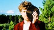 Ein junger Mann schaut in die Kamera und hält dabei das linke Auge mit einer Tonband-Rolle bedeckt. Im Hintergrund ein Wald und blauer Himmel. © Horizonte Festival Göttinger Symphonie Orchester Foto: Finn Waldherr