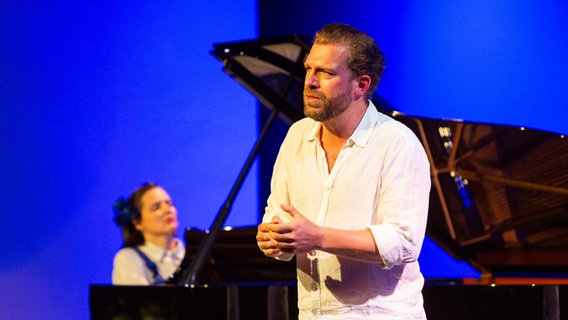 Ein Mann auf einer Bühne, in blaues Licht getaucht. Im Hintergrund eine Pianistin am Flügel. © Jörg Modrow Foto: Jörg Modrow