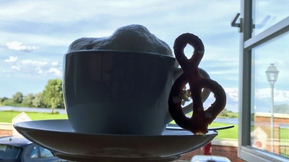 Neben einer Tasse Kaffee mit einer Milchhaube lehnt ein Stück Gebäck in Form eines Notenschlüssels. © NDR Foto: Raliza Nikolov