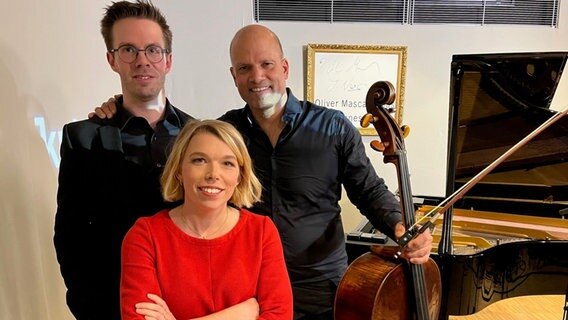 Zwei große Männer stehen hinter einer Frau, die ein rotes Kleid trägt. Die drei Personen stehen neben einem Flügel und einem Cello. © NDR / Claus Röck Foto: Claus Röck