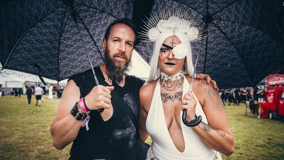 Ein Mann mit Bart in Schwarz und eine Frau in weiß, beide mit schwarzen Sonnenschirmen, auf dem Festival M'era Luna 2023 © NDR Foto: Benjamin Hüllenkremer