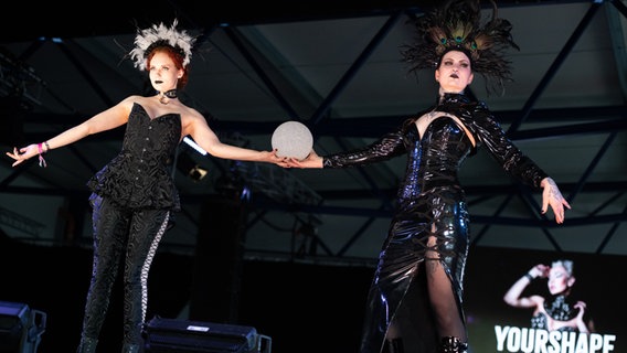 Zwei Frauen zeigen ihre Outfits und halten eine Kugel bei einer Fashion Show im Flugzeughangar beim M'era Luna Festival 2023 © NDR Foto: Benjamin Hüllenkremer