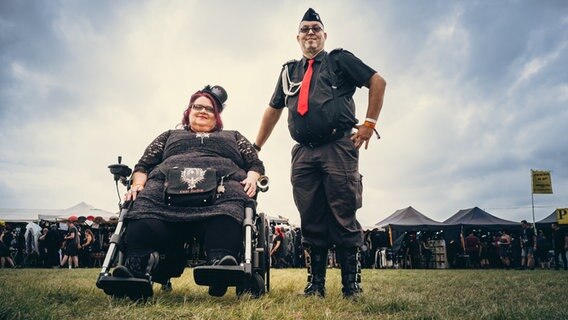 Eine Frau in schwarz mit Zylinder im Rollstull, daneben ein Mann mit Barrett - Fans beim Festival M'era Luna 2023 © NDR Foto: Benjamin Hüllenkremer