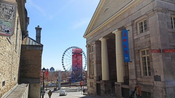 In mitten zweier alter Gebäude in der Innenstadt Montréals sieht man ein Riesenrad im Hintergrund. © Eva Schramm / NDR Foto: Eva Schramm