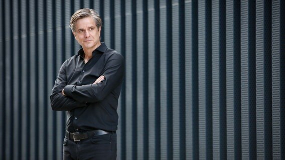 Ein Mann steht mit verschränkten Armen vor einer grauen Wand. © picture alliance/dpa/Peppermint Park | Nadja Mahjoub Fotografie Foto: Nadja Mahjoub