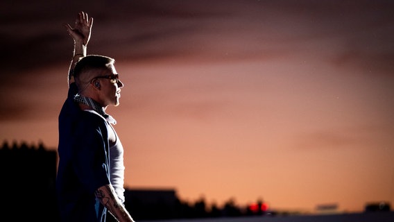 Der Rapper Macklemore steht mit erhobenen Armen auf einer Bühne im Sonnenuntergang © picture alliance Foto: Nuno Cruz