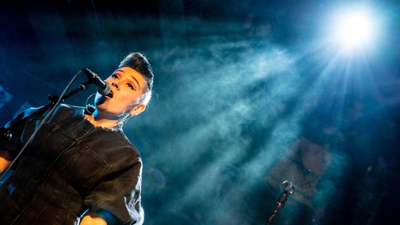 Eine Sängerin steht am Mikrofon im Bühnennebel und wird von einem Scheinwerfer angestrahlt. © IMAGO / xim.gs 