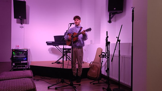 Ein junger Mann steht mit einer Gitarre in einem lila ausgeleuchteten Raum. © NDR / Svenja Estner Foto: Svenja Estner