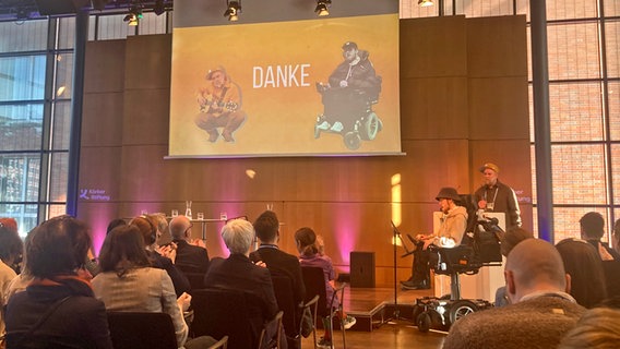 Menschen sitzen vor einer Bühne, ein junger Mann sitzt im Rollstuhl, sein Foto wird auf eine Leinwand projiziert © NDR Foto: Marcus Stäbler