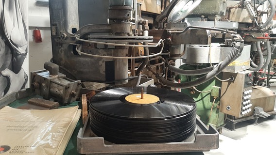 Ein Stapel fertiger Schallplatten liegt vor einer großen Plattenpresse. Ein Stapel fertiger Schallplatten liegt vor einer großen Plattenpresse. © Dudda Foto: Alexa Dudda