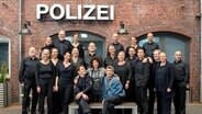 Das NDR Vokalensemble steht mit Darstellern der Serie "Großstadtrevier" vor einer Wand mit der Aufschrift "Polizei" bei Dreharbeiten zur 500. Folge © ARD/Thorsten Jander/Montage Foto: THorsten Jander