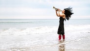 Eine Frau mit wehenden schwarzen Haaren und roten Gummistiefeln steht am Meer und spielt Trompete. © photocase.de Foto: Nordreisender