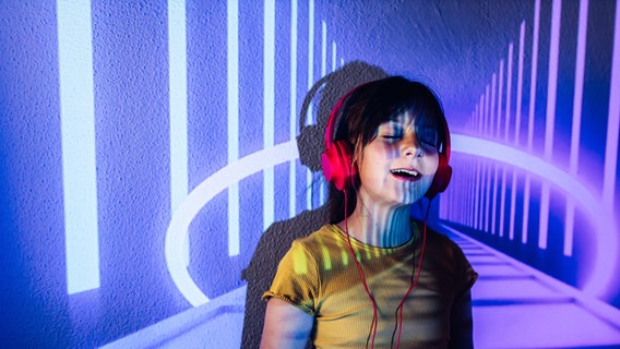 Ein Mädchen mit Kopfhörern steht vor einer lilafarbenen Leinwand © picture alliance / Shotshop | Addictive Stock 