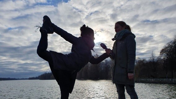 Balletttänzerin Michèle Seydoux zeigt im Sonnenschein am Maschsee eine Ballettfigur und hebt dabei ein Bein nach hinten und hält den Fuß mit der Hand fest. NDR-Reporterin Andrea Schwyzer steht daneben mit einem NDR Mikrofon. © Andrea Schwyzer / NDR Foto: Andrea Schwyzer