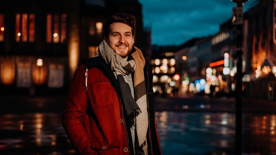 Jakob Lübke lächelt mit dickem Schal und Jacke in die Kamera. Hinter ihm leuchten viele Lichter einer Großstadt. © Birgitt Schütte / NDR 