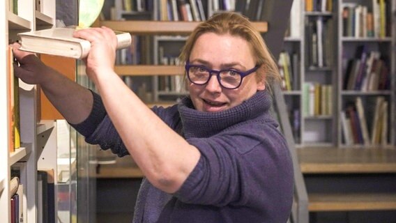 Katharina Hanschel stellt ein Buch zurück in die Regale, dabei schaut sie in die Kamera. © Juliane Voigt / NDR Foto: Juliane Voigt