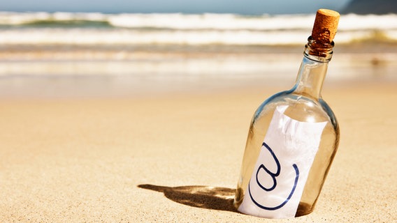 Eine Flaschenpost mit eine Zettel und @-Zeichen steckt halb im Sand an einem Strand. © Don Bayley Foto: RapidEye