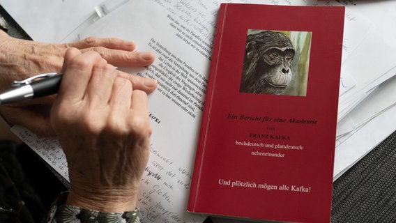 Ein rotes Buch mit Affenkopfzeichnung und Papiere liegen auf einem Tisch. Eine Hand hält einen Kugelschreiber. © NDR Foto: Lornz Lorenzen