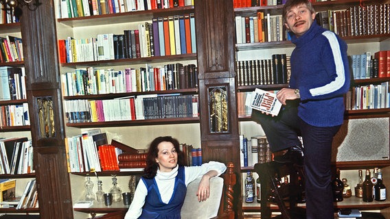 Der deutsche Schauspieler Uwe Friedrichsen steht auf einem Stuhl vor seinem Bücherregal. Seine Verlobte Winka Wulff sitzt daneben in einem Sessel. © dpa - Bildarchiv 