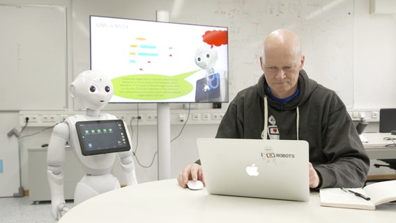 Der KI-Experte Thomas Sievers sitzt am Laptop, neben ihm steht der Roboter Pepper. © NDR Foto: Jorrit Groth