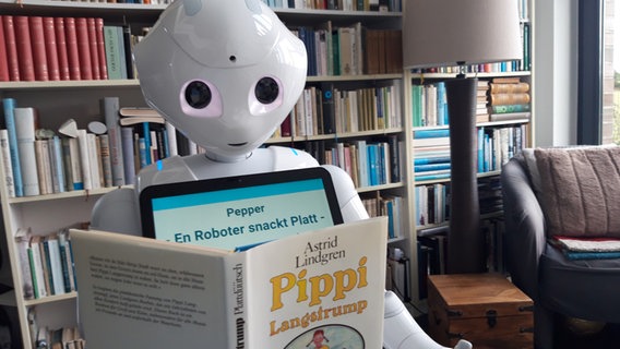 Der Roboter Pepper blick auf ein aufgestelltes Buch "Pippi Langstrumpf" von Astrid Lindgren. Hinter ihm ist ein großes Bücherregal. © NDR/Thomas Sievert Foto: Thomas Sievert