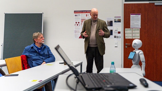 Ein Mann (Informatiker Thomas Sievers) hält einen Vortrag in einem Seminarraum, neben ihm steht ein Roboter. © NDR/Lornz Lorenzen Foto: Lornz Lorenzen