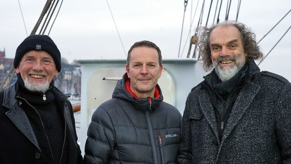 Drei Mitglieder der Band Santiano Björn Both (l.), Axel Stosberg (m.) und Hans-Timm Hinrichsen (r.) stehen auf einem Segelschiff und lächeln in die Kamera. © NDR 