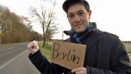 Gerrit Hoss steht auf einer Straße und hält ein Pappschild in die Höhe auf dem Berlin steht, dazu hebt er den "Tramper-Daumen". © NDR Foto: Gerrit Hoss