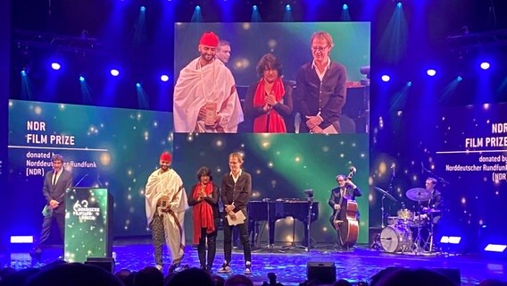 Auf der Bühne im Theater Lübeck wurde der NDR Filmpreis vergeben an den Regisseur Khadar Ayderus Ahmet neben zwei Laudatoren. © Linda Ebener Foto: Linda Ebener