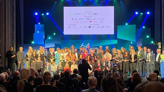 Im Theater Lübeck stehen alle Preisträgerinnen und Preisträger, sowie alle Jurymitglieder und Mitwirkenden auf der Bühne vor einem blau-grünen Hintergrund. © Linda Ebener / NDR Foto: Linda Ebener