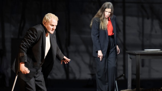 Die Darsteller Devid Striesow und Julia Wieninger stehen auf einer dunklen Bühne, er hält sich mit einer Krücke aufrecht. © Monika Ritterhaus 