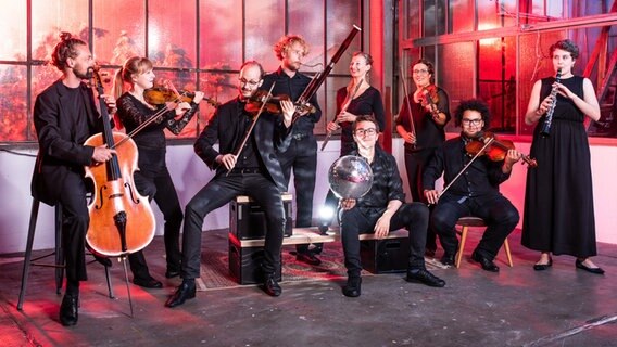 Die Mitglieder des Orchesters im Treppenhaus mit ihren Instrumenten in einer farbig ausgeleuchteten Fabrikhalle ©  Moritz Küstner Foto:  Moritz Küstner