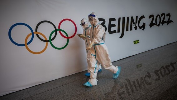 Helfer in kompletter Schutzmontur gehen an den Olympischen Ringen und dem Schriftzug "Beijing 2022" vorbei. © picture alliance/dpa Foto: Michael Kappeler